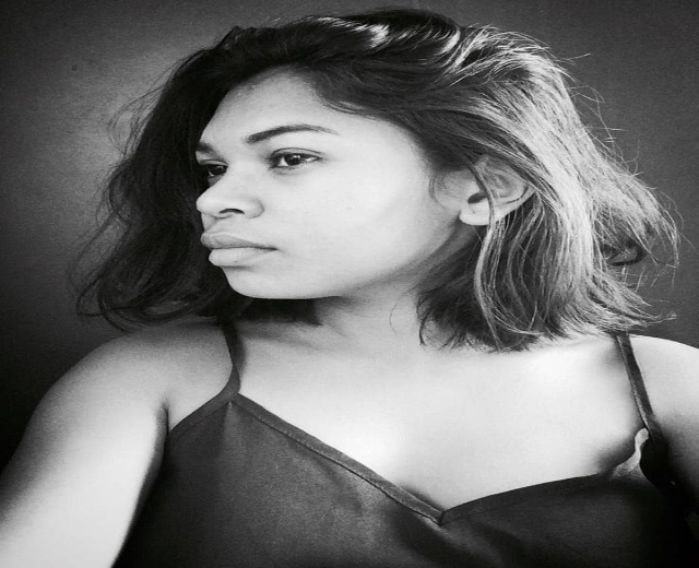 Eine selbstbewusste Frau in einem schwarzen Kleid posiert in einem Schwarz-Weiß-Foto, steht mit einer selbstbewussten Haltung vor einer Wand, ihr glatter Bob und dezentes Make-up unterstreichen ihren entschlossenen Ausdruck.