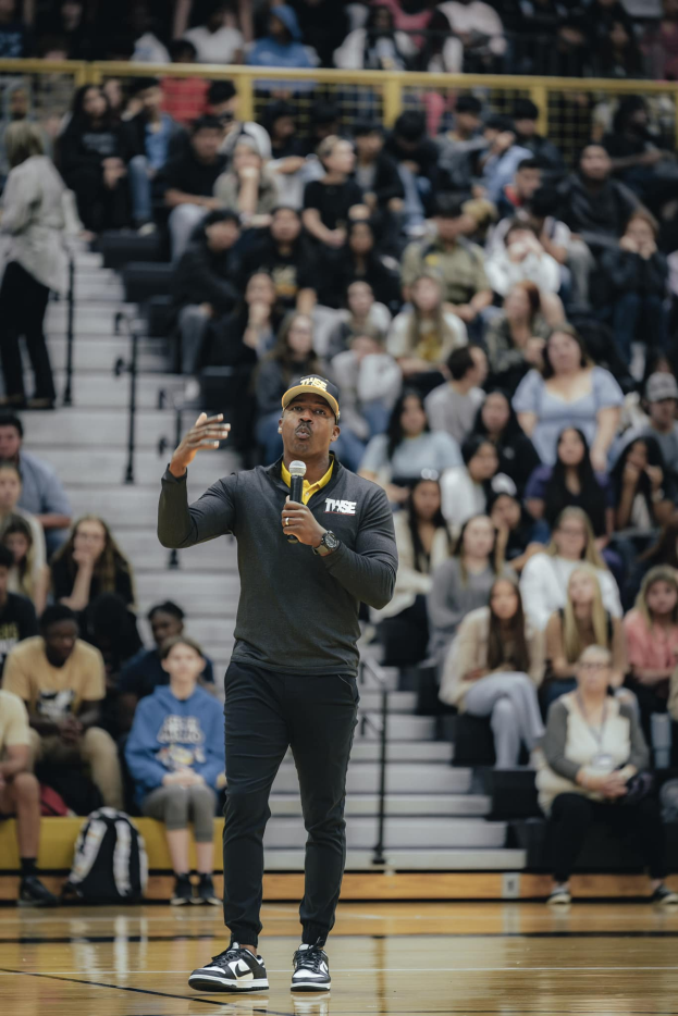 Ein Mann in einer Mütze mit einem Mikrofon auf einem Basketballfeld stehend, mit einer Gruppe von Menschen auf der Treppe und Taschen auf dem Boden dahinter.