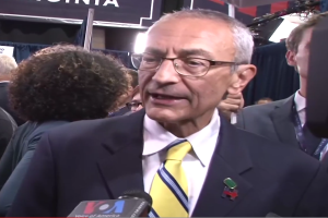 Senator Chuck Schumer in Anzug und Krawatte spricht zu Reportern an einem Podium, umgeben von Menschen in Mänteln und ID-Karten, mit Schildern im Hintergrund, auf denen "Sen. Schumer sagt, er sei 'kein Demokrat'".