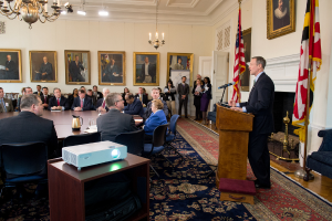 Ein Mann spricht an einem Rednerpult auf der rechten Seite, mit einigen Zuhörern auf der linken Seite, einem Projektor auf einem Tisch, Fotorahmen an der Wand, zwei Flaggen auf der rechten Seite und Lampen mit einem Kronleuchter oben.