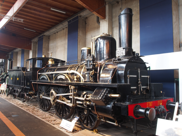 Alte Dampflokomotive im Museum ausgestellt, umgeben von Eisenbahnschienen, Informationsschildern und anderen Ausstellungsstücken, mit einer Wand und Deckenbeleuchtung im Hintergrund.