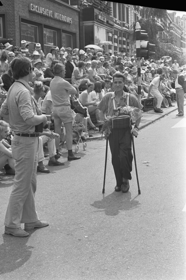 Ein Mann mit einem Stock geht die Straße entlang an einer Gruppe von sitzenden und stehenden Menschen vorbei, mit einer Ampel, Gebäuden und Bäumen im Hintergrund in Schwarz-Weiß.