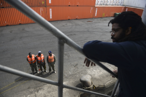 Ein Mann in Freizeitkleidung steht auf einem Metallgelönder in der Nähe einer Gruppe von Menschen mit orangen Jacken und Kappen, mit Containern und einer Wand im Hintergrund am Hafen von Los Angeles.