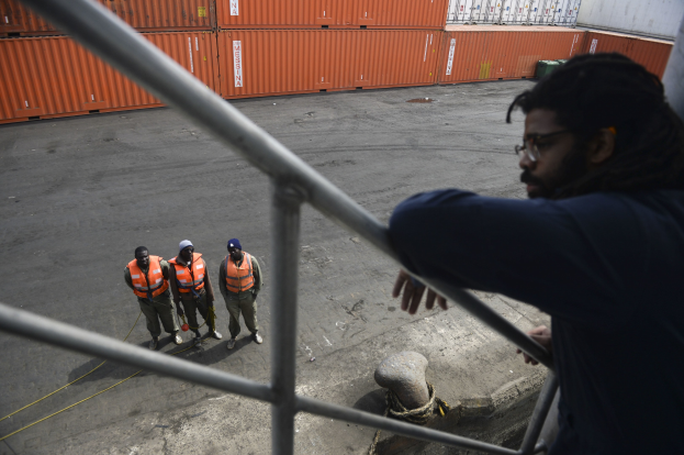 Ein Mann in Freizeitkleidung steht auf einem Metallgelönder in der Nähe einer Gruppe von Menschen mit orangen Jacken und Kappen, mit Containern und einer Wand im Hintergrund am Hafen von Los Angeles.