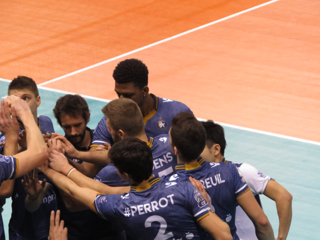 Eine Gruppe von Männern in blauen 'Ligue 1'-T-Shirts feiert auf einem Volleyballfeld, einige halten die Hände siegreich in die Luft.