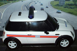 Ein weißer Polizeiwagen steht im Vordergrund, hinter ihm bewegen sich mehrere Fahrzeuge auf der Straße, umgeben von Gras und Bäumen zu beiden Seiten in einer ländlichen Umgebung.