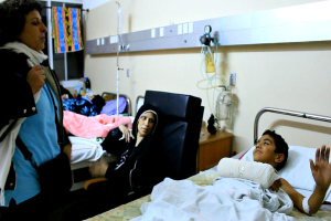 Ein Krankenhauszimmer mit mehreren Betten, von denen jedes von einem Patienten belegt ist, und eine Frau, die in der Nähe eines der Betten steht und eine Tasche trägt.