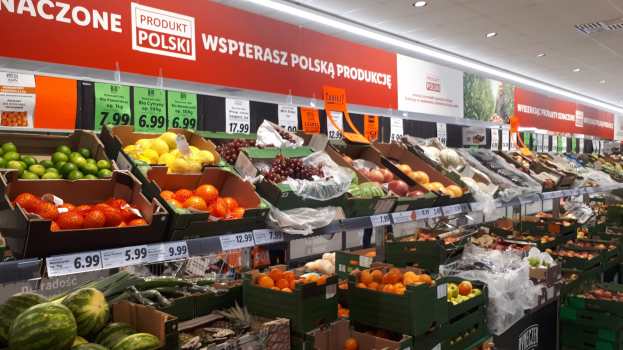 Ein Geschäft mit verschiedenen Obst- und Gemüsesorten in Kartonschachteln, jeweils mit Preisschildern, beleuchtet von Deckenlampen und dekoriert mit Text- und Bildtafeln an den Wänden.