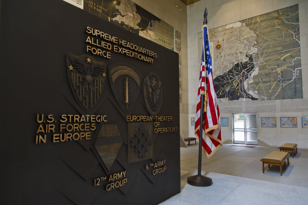 Eingang zum Hauptquartier der US-Strategischen Luftstreitkräfte in Europa, mit einer amerikanischen Flagge, einer Wand mit Text und Logos, Bänken, einer Tafel, Fotorahmen, beleuchteter Decke und einer Tür im Hintergrund.
