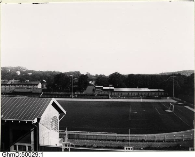 Schwarzes und weißes Foto eines Fußballfeldes mit einer Hütte links, einem umliegenden Zaun, zentralen Pfosten und Bäumen mit Himmel im Hintergrund, beschriftet mit "Gillingham Football Club, 1960er Jahre" unten.