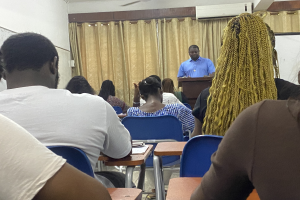 Ein als Gesundheits- und Sozialminister identifizierter Mann steht an einem Rednerpult und spricht vor einer Klasse sitzender Personen, wobei Bücher und Gegenstände auf Tischen, ein Vorhang und eine Klimaanlage im Hintergrund zu sehen sind.