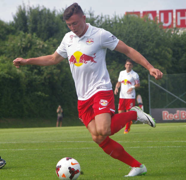 Ein Profifußballer kickt einen Ball auf einem Rasenfeld mit Bäumen und einem klaren blauen Himmel im Hintergrund, in der Nähe eines Schildes mit der Aufschrift "RB Leipzig."