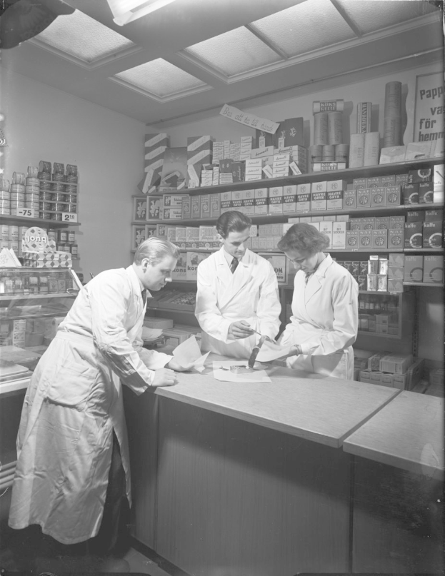 Drei Männer in weißen Kitteln an einer Apotheken-Theke mit Papieren und Gegenständen, auf einem Schwarz-Weiß-Bild mit Regalen im Hintergrund.