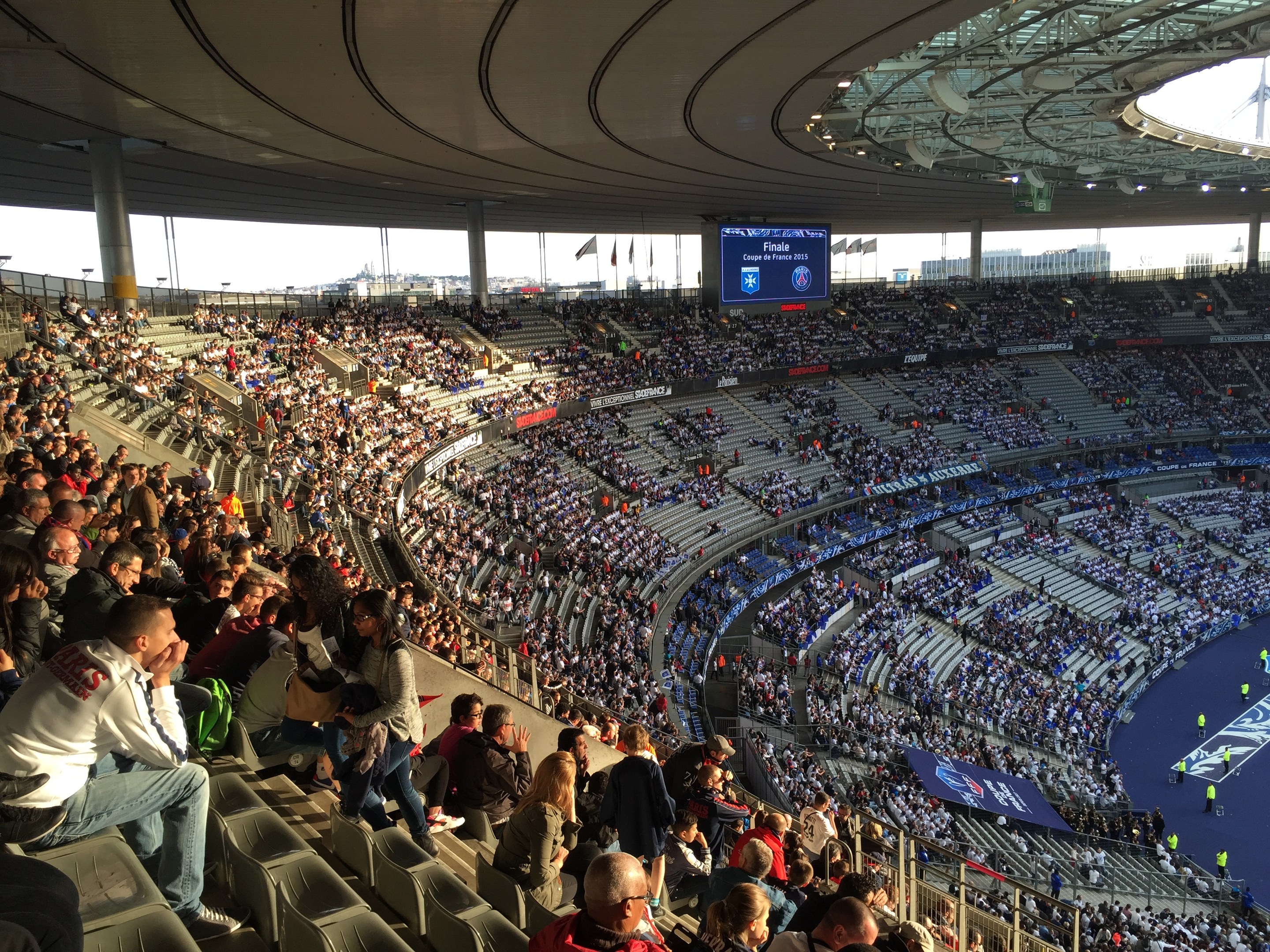 Große Menschenmenge in einem Stadion bei einem Fußballspiel mit einer Bühne, Fahnen und einem Bildschirm im Hintergrund, identifiziert als Allianz Arena in München, Deutschland.