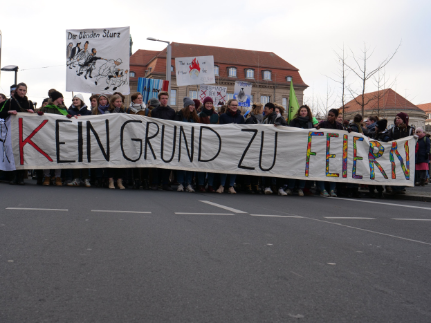 Demonstranten mit einem Transparent mit der Aufschrift 'Kein Grund zur Feier' gegen deutsche Sparmaßnahmen, mit Straßeninfrastruktur und Gebäuden im Hintergrund bei klarem Himmel.