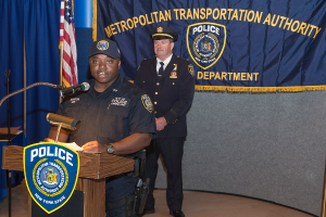 Zwei Polizisten stehen vor einem Rednerpult und einer Tafel mit Symbolen und Text, wobei einer der Polizisten Papiere hält und in ein Mikrofon spricht, eine Fahne, ein Stöß, ein Banner, ein Tisch und ein Stand sind ebenfalls zu sehen.
