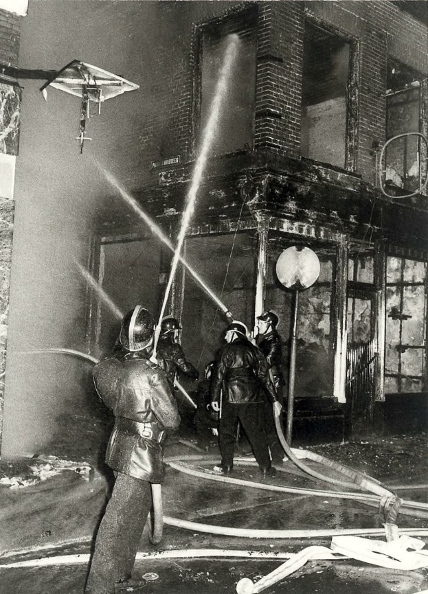 Schwarzes und weißes Bild von Feuerwehrmännern in Helmen mit Rohren, die vor einem feuerbeschädigten Gebäude stehen.
