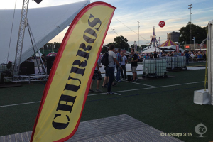 Eine lebhafte Faschingszene mit Zelten, einem zentralen Mast, Menschen, Bäumen, Strommasten, Straßenbahnen auf der Straße und einem eisernen Stand auf der linken Seite unter einem klaren Himmel.