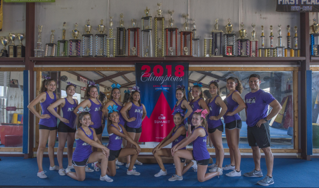 Gruppe von Cheerleadern in lila T-Shirts und schwarzen Shorts, die mit einer Vitrine voller Trophäen posen, einige stehen, andere knien, mit einem "2018 Summit Cheerleading Champions"-Banner im Hintergrund.