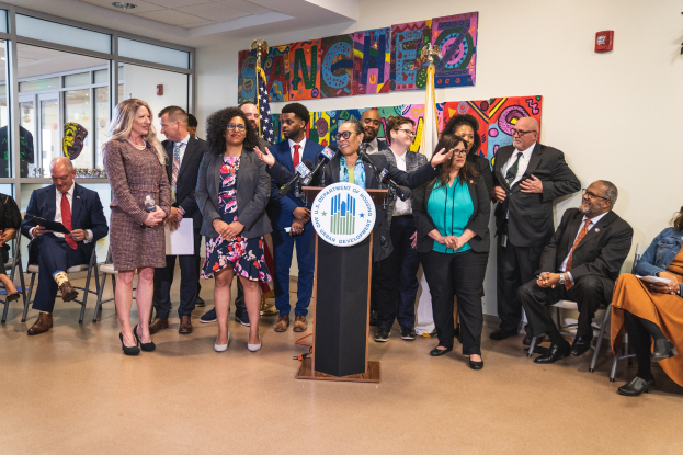 Gruppe von Menschen vor einem Rednerpult mit einem Mikrofon und einer Tafel, auf der "Sancho Stadtrat joins City Council" steht, mit sitzenden und stehenden Teilnehmern, Flaggen, einem Feuerlöscher und Deckenleuchten im Hintergrund.