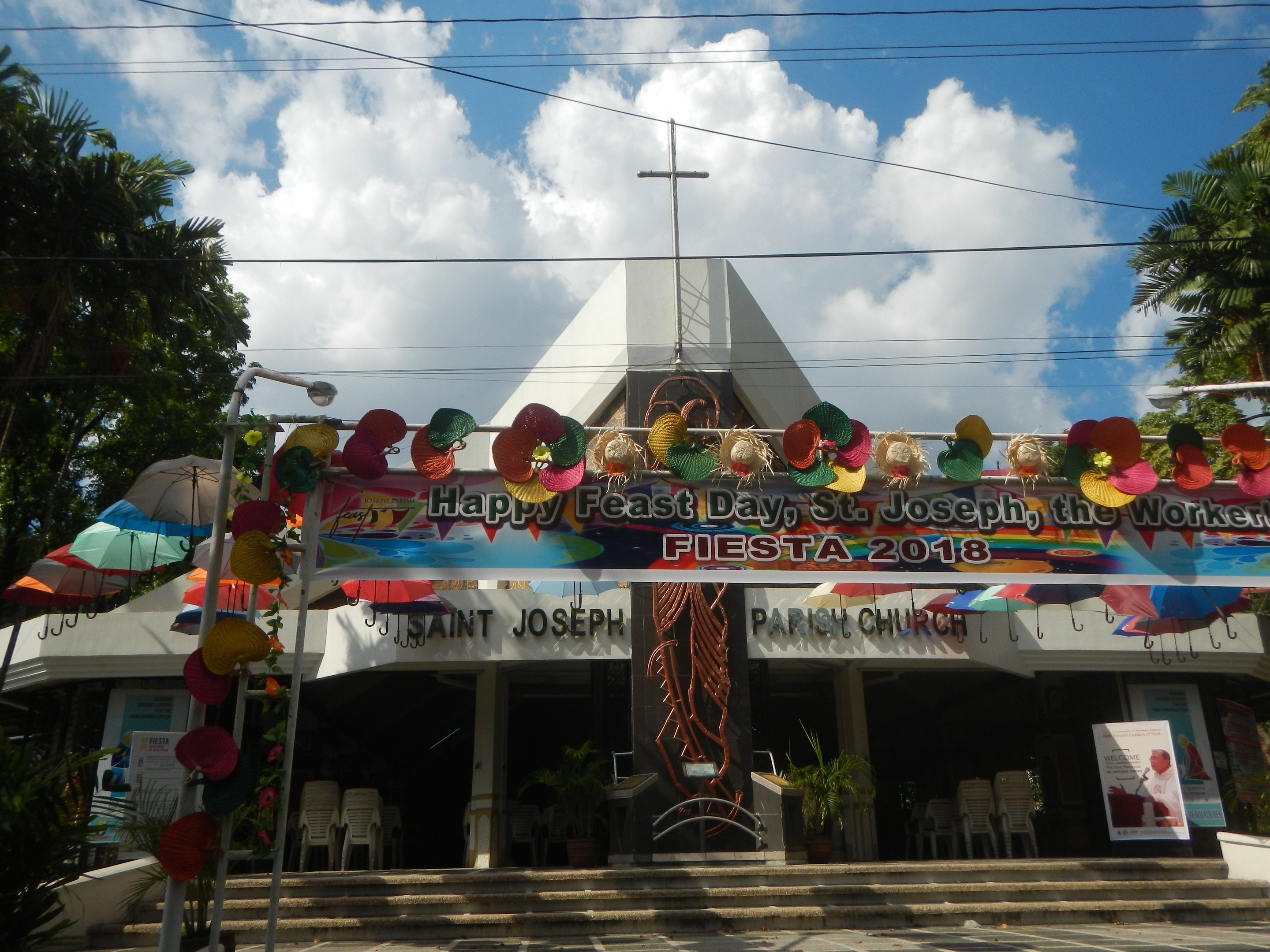 Ein Gebäude mit einem "Frohes Fest St. Josef der Arbeiter"-Schild, Treppe zum Eingang, ein Bogen mit hängenden Dekorationen, Außensitzbereich mit Tischen und Stühlen, Topfpflanzen, Bäume auf beiden Seiten und ein bewölkter Himmel.