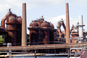 Ein großes Fabrikgebäude mit zahlreichen Rohren und Schornsteinen, einer Brücke, Masten, Lampen, Bäumen und einem wolkenverhangenen Himmel im Hintergrund.