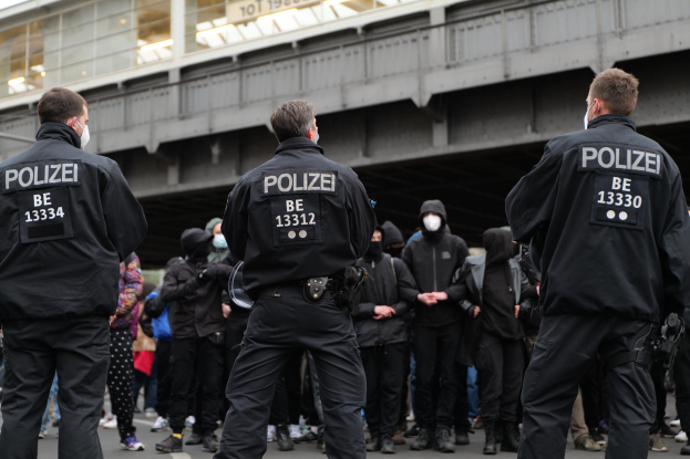 Eine Gruppe von Polizisten in Uniform steht vor einer Menge maskierter Individuals in Schwarz, mit einer Stadtbrücke und einem Gebäude im Hintergrund, während einer Demonstration.
