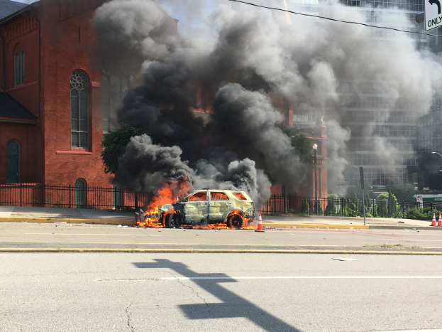 Ein Auto brennt auf der Straßenseite mit Rauchwolken, umgeben von Gebäuden, Bäumen und anderen urbanen Elementen unter einem klaren Himmel.