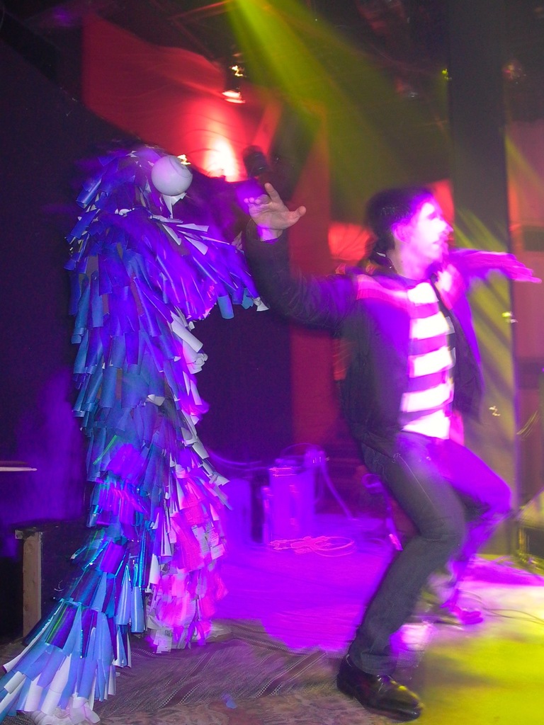 Leute tanzen auf der Bühne, wobei eine Person links ein elegantes Kostüm trägt und Scheinwerfer oben sichtbar sind.