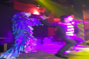 Leute tanzen auf der Bühne, wobei eine Person links ein elegantes Kostüm trägt und Scheinwerfer oben sichtbar sind.