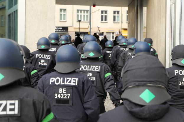 Gruppe von Polizisten in Uniform und Helmen vor einem Gebäude mit einigen Menschen und Gebäuden im Hintergrund.