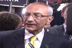 Ein Mann im Anzug und mit Krawatte, der eine Brille trägt, spricht zu Reportern mit einem Mikrofonständer vor ihm, umgeben von einer Gruppe von Menschen, mit Lichtern und einer Tafel im Hintergrund.