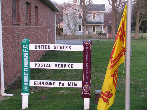 Ein grasbewachsener Bereich mit einem Mast in der Mitte, der Flaggen und ein Schild der 'United States Postal Service' trägt, ein Gebäude mit Fenstern links und Bäume mit einem klaren blauen Himmel im Hintergrund.
