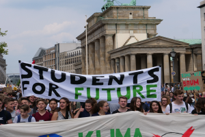 Eine Gruppe von Schülern marschiert in Berlin, die ein buntes Banner mit der Aufschrift "Students for Future" tragen, vor dem Hintergrund von Gebäuden, Bäumen und Himmel.