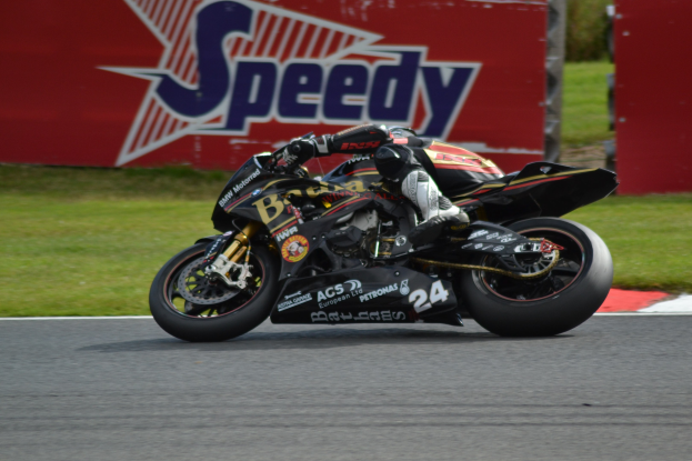 Eine Person in Helm und Schuhwerk fährt ein Motorrad auf einer Rennstrecke, umgeben von Gras, mit einer Tafel im Hintergrund, auf der Text zu sehen ist.