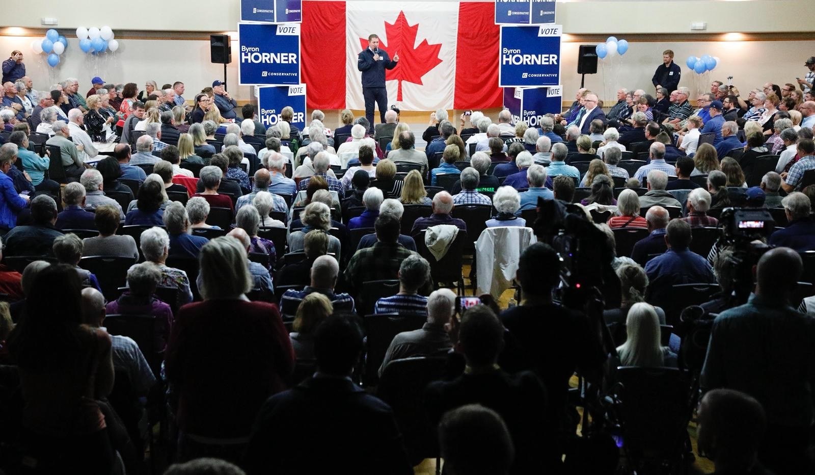 Eine große Menschenmenge sitzt auf Stühlen, die einem kanadischen Flagge gegenüberstehen, mit einem Mann auf einem Podium, der ein Mikrofon hält, umgeben von Bannern, Luftballons, Lautsprechern und einer Wand im Hintergrund.