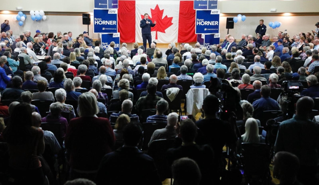 Eine große Menschenmenge sitzt auf Stühlen, die einem kanadischen Flagge gegenüberstehen, mit einem Mann auf einem Podium, der ein Mikrofon hält, umgeben von Bannern, Luftballons, Lautsprechern und einer Wand im Hintergrund.