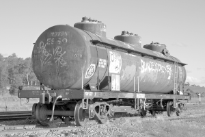 Ein Güterzug auf Eisenbahnschienen mit Bäumen, einem Pfahl und einem Himmel im Hintergrund und Steinen, Schienen und Gras im Vordergrund.