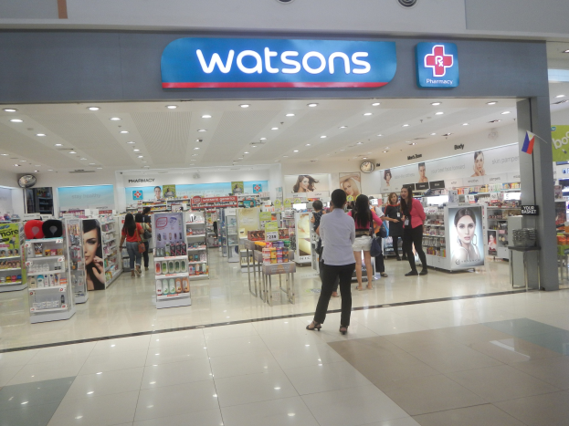 Watsons Apotheken-Interieur in einem Einkaufszentrum mit Menschen, Regalen, Schildern, einer Wanduhr und Deckenbeleuchtung.