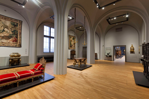 Ein geräumiger Raum im Kunsthistorischen Museum in Wien, Österreich, mit einem Sofa mit Kissen, Tischen mit verschiedenen Gegenständen, Säulen, Fenstern und Deckenlampen, sowie zahlreichen paintings an den Wänden.