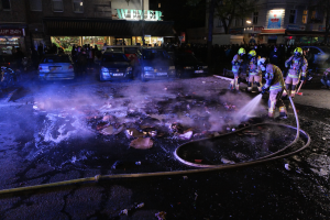 Eine Gruppe von Feuerwehrleuten in Helmen, die Rohre halten, steht um ein Straßenfeuer herum mit Fahrzeugen in der Nähe, Gebäuden, Schildern, Laternenpfählen, Bäumen und einem dunklen Himmel im Hintergrund.