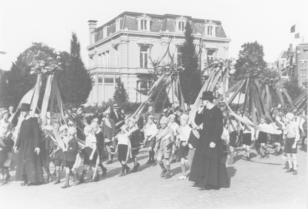 Schwarzes und weißes Bild einer Gruppe von Menschen, die vor einem Gebäude die Straße entlanggehen, einige halten Fahnen, tragen Hüte, Bäume, Gebäude und einen Maibaum im Hintergrund.