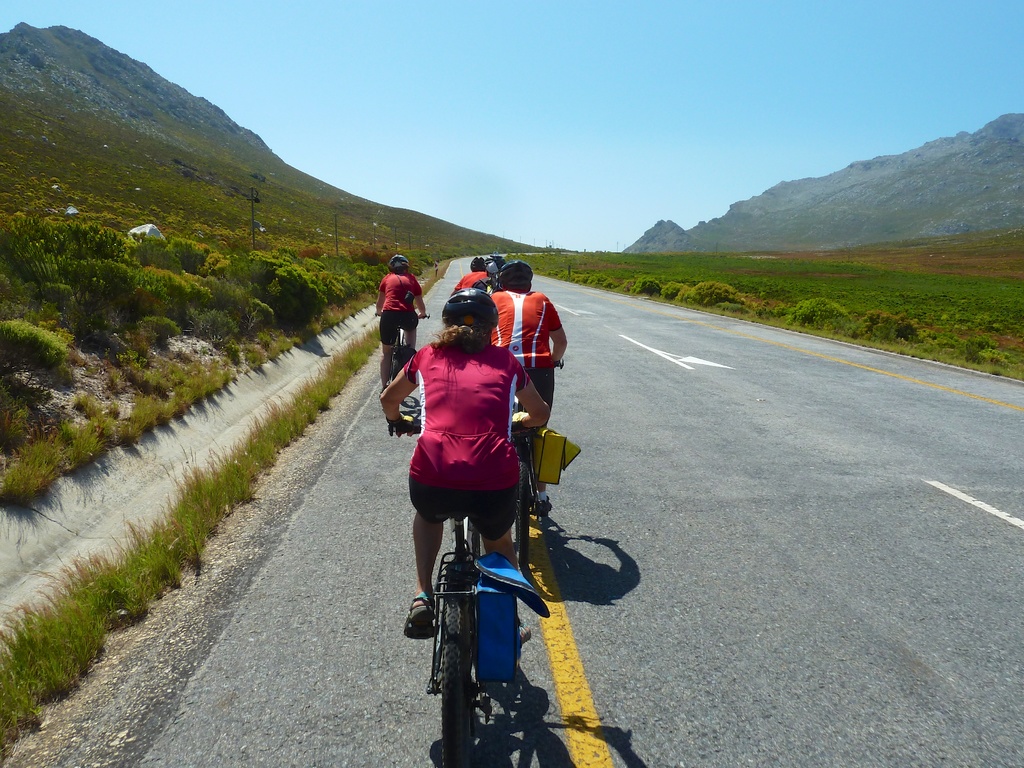 Eine Gruppe von Menschen mit Helmen auf Fahrrädern auf einer Straße, mit Pflanzen, Gras, Masten, Bergen und einem bewölktem Himmel im Hintergrund.