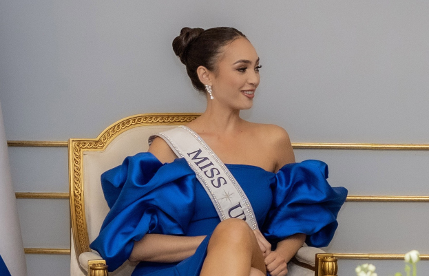 Frau in einem blauen Kleid mit einer "Miss Universe 2019" Schärpe, die auf einem Stuhl sitzend und lachend umgeben von Blumen mit einer Wand im Hintergrund.