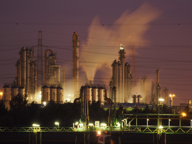 Ölraffinerie bei Nacht mit aufsteigendem Rauch, beleuchtet von umliegenden Lichtern, vor einem sichtbaren Himmel und mit einem Wasserzeichen.