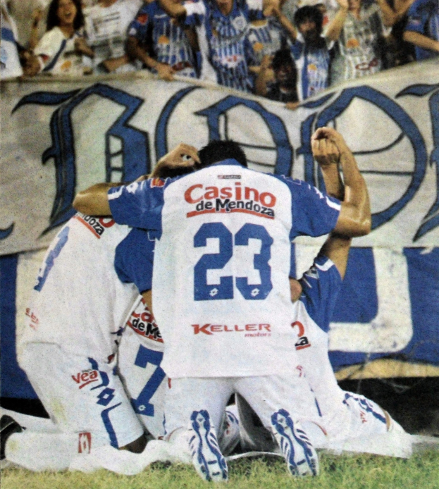 Gruppe von Fußballspielern, die ein Tor feiern, auf dem Feld mit einer jubelnden Menge und einem Banner im Hintergrund.