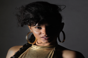 Eine Frau in einem goldenen Kleid und großen Creolen steht selbstbewusst vor einer Wand, ihr glatter Bob und natürliches Make-up unterstreichen ihren entschlossenen Gesichtsausdruck, während sie direkt in die Kamera schaut.