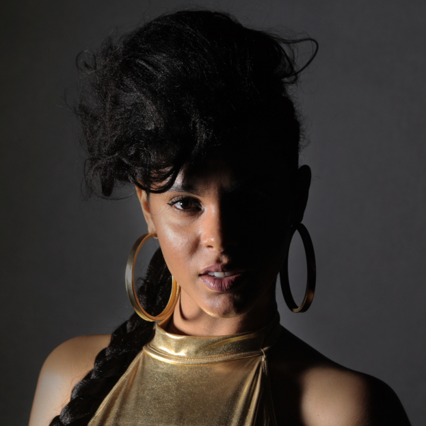 Eine Frau in einem goldenen Kleid und großen Creolen steht selbstbewusst vor einer Wand, ihr glatter Bob und natürliches Make-up unterstreichen ihren entschlossenen Gesichtsausdruck, während sie direkt in die Kamera schaut.