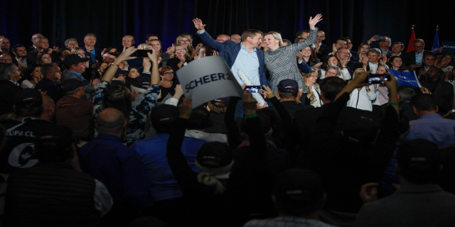 Ein Mann und eine Frau stehen vor einer Menge, einige halten Schilder und Handys, sowie Fahnen und einen Vorhang im Hintergrund.