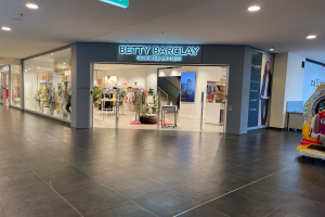 Das Innere eines Betty Barclay-Ladens in einem Einkaufszentrum, das Schaufensterpuppen in Kleidern, hängende Kleidung, Tische, Pflanzen und andere Gegenstände zeigt, mit einem Spielzeugfahrzeug auf der rechten Seite und einer Tafel mit der Aufschrift "Betty Barclay Selected Brands" oben.